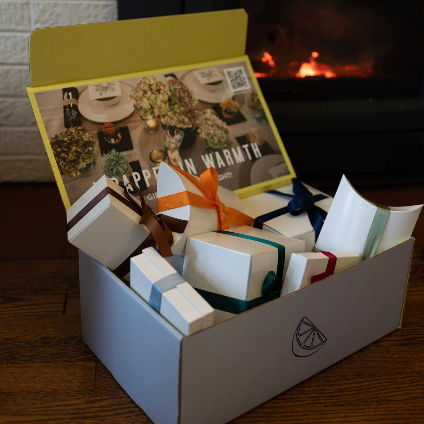 Gift box with wrapped presents and a card on a wooden surface with a fireplace in the background